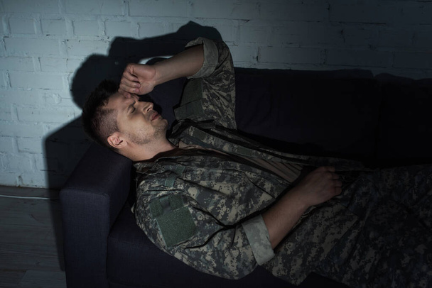 Στεναχωρημένος στρατιωτικός με στολή που υποφέρει από μετατραυματικό στρες ενώ είναι ξαπλωμένος στον καναπέ.  - Φωτογραφία, εικόνα