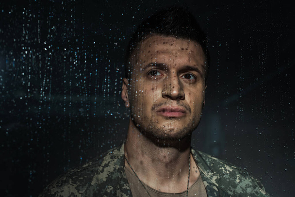 triste militar en uniforme militar mirando por la ventana lluviosa mientras sufre de trastorno de estrés postraumático  - Foto, Imagen