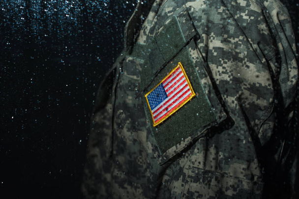 cropped view of American serviceman in military uniform with flag standing behind rainy window  - 写真・画像