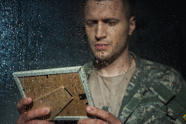 triste militar mirando el marco de fotos y tener flashbacks mientras está de pie junto a la ventana con gotas de lluvia  - Foto, Imagen