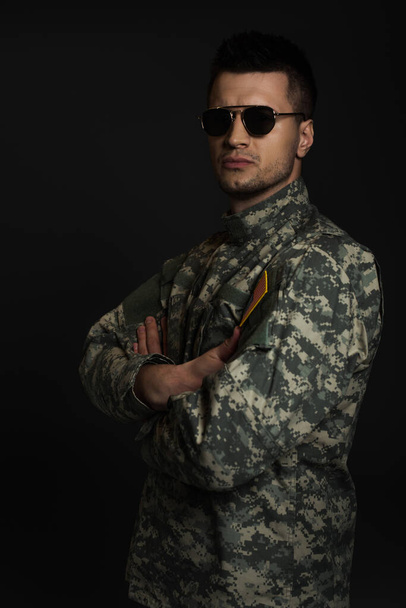 soldado en uniforme de camuflaje y gafas de sol mirando a la cámara mientras está de pie con los brazos cruzados aislados en negro  - Foto, Imagen