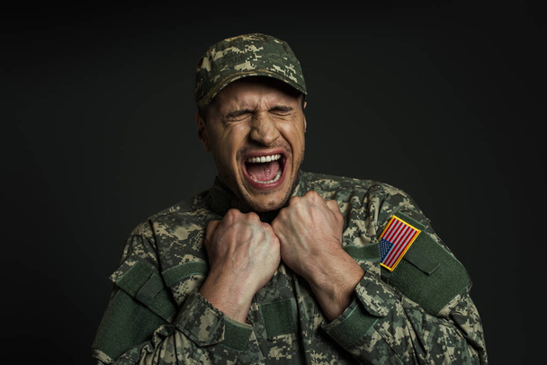 soldado estresado en uniforme con bandera de EE.UU. gritando mientras sufría de ataques de pánico aislados en negro  - Foto, imagen