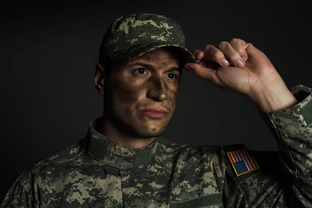 military man with dirt on face standing in uniform and adjusting cap isolated on grey  - Photo, Image