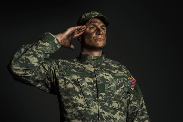 patriotický voják ve vojenské uniformě salutuje, zatímco stojí izolovaný na šedi  - Fotografie, Obrázek