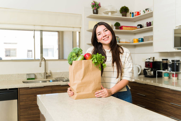 Привлекательная счастливая женщина с вегетарианским образом жизни на кухне готова готовить здоровую пищу с овощами после похода в супермаркет - Фото, изображение