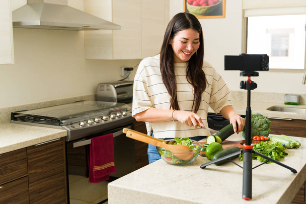 Glückliche Influencerin filmt ein Kochvideo, während sie ein veganes Rezept zubereitet und mit der Kamera in ihrer Küche filmt - Foto, Bild