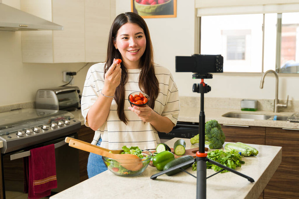 Feliz influencer caucásico sonriendo mientras filma un video de blog y cocina una receta vegana con verduras y frutas orgánicas - Foto, imagen