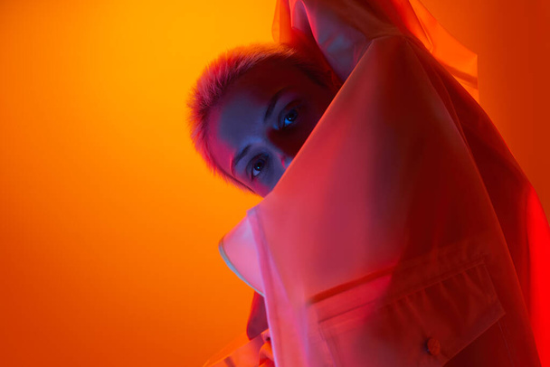 Seitenansicht des selbstbewussten jungen weiblichen Modells mit kurzen blonden Haaren im transparenten Regenmantel, das in Neonlichter vor orangefarbenem Hintergrund in die Kamera blickt - Foto, Bild