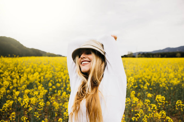 Schöne junge blonde, fröhliche Frau mit Hut inmitten eines Feldes mit blühenden gelben Rapsblüten - Foto, Bild