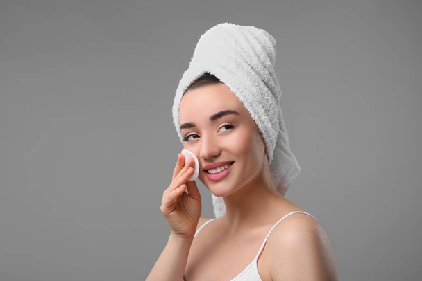 Beautiful woman in terry towel removing makeup with cotton pad on gray background - Фото, изображение
