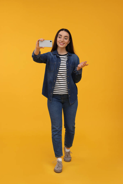 Smiling young woman taking selfie with smartphone on yellow background - Foto, Bild