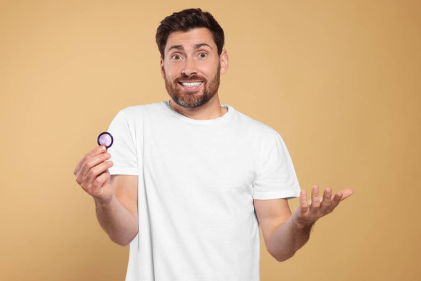 Confused man holding condom on beige background. Safe sex - 写真・画像