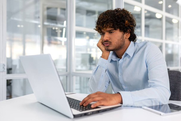 Upset thinking businessman working with laptop inside modern office, man frustrated solving difficult decision at workplace, hispanic man with curly hair serious in shirt. - Foto, immagini
