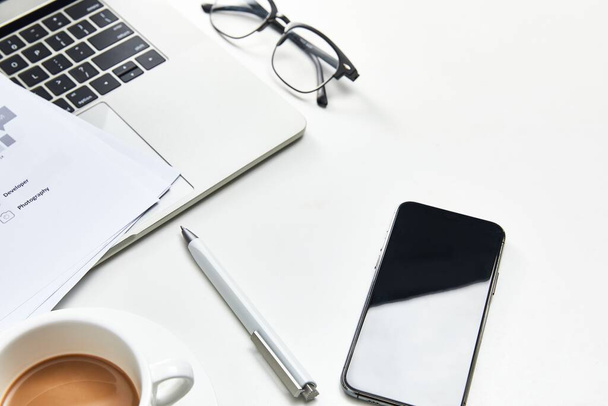 Μια εναέρια λήψη ενός τηλεφώνου, γυαλιά, στυλό, laptop, μερικά αρχεία και ένα φλιτζάνι καφέ σε λευκό φόντο - Φωτογραφία, εικόνα