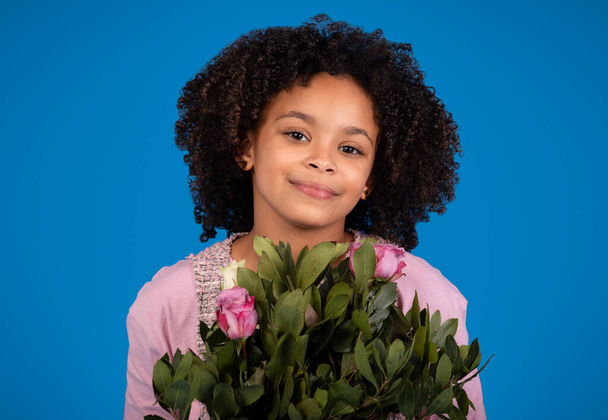 Positive african american little girl with bouquet of flowers enjoy birthday, isolated on blue background, studio, close up. Holiday celebration, congratulation and children emotions - Foto, Imagem