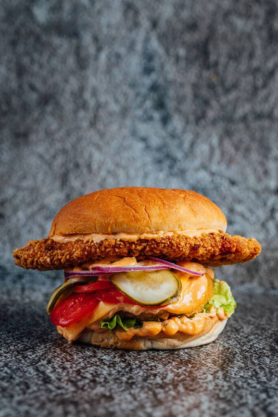 A vertical closeup shot of a large fried chicken cheeseburger with vegetables on a gray surface - Фото, изображение