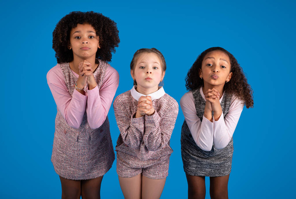 Uluslararası küçük kızlar dua etmek için jest yapıyor, mavi arka planda, stüdyoda izole edilmiş kameraya bakıyorlar. Umut, hayal, okulda büyük bir eğitim arzusu, çocuk duyguları. - Fotoğraf, Görsel