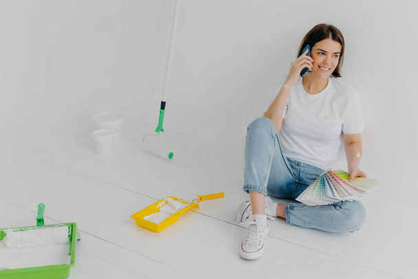 Glad brunette woman in casual clothes, holds color samples, chooses appropriate tone for room, asks for advice in friend, calls via smartphone, poses in new bought apartment, isolated on white wall - Fotoğraf, Görsel