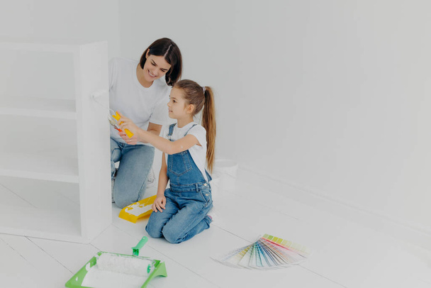 Little girl learns how to paint with roller, stands on knees, her mother tells how to do it correctly, pose in spacious white room, choose color from palette samples. Home makeover, decoration concept - Foto, imagen