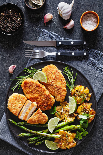 fried breaded chicken breasts with roast asparagus, smashed potatoes and lime on black plate on dark concrete table with cutlery, horizontal view from above - Photo, Image