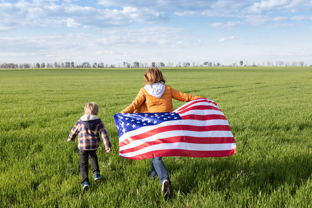 Kadın ve bir çocuk neşeyle yeşil bir çayırda koşuyor, arkalarında Amerikan bayrağı dalgalanıyor. Amerikalı olmaktan gurur duyuyorum. Özgürlüğün ulusal sembolü, Amerika Birleşik Devletleri 'nin bağımsızlık günü - Fotoğraf, Görsel