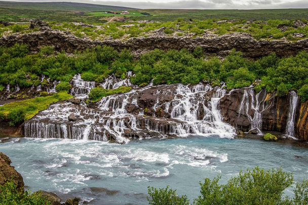 The beautiful waterfall flowing over the rocks in Iceland with lush trees in the background - Fotoğraf, Görsel
