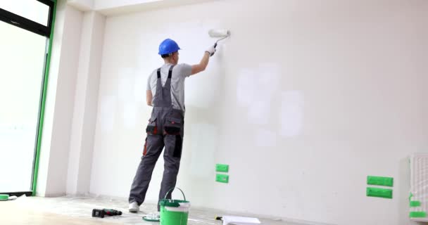 Foreman schildert muur in het wit met behulp van roller. Werknemer in helm en professionele uniform helpt eigenaren renovatie doen in appartement achteraanzicht - Video