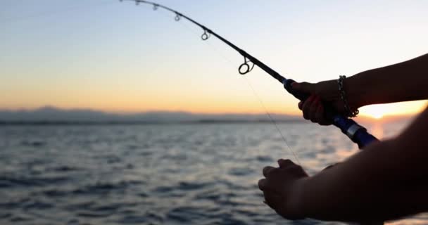 Vrouwelijke hand houdt vishengel vast. Vrouw vist op zeilmotorboot bij zonsondergang. Actieve recreatie en hobby slow motion - Video