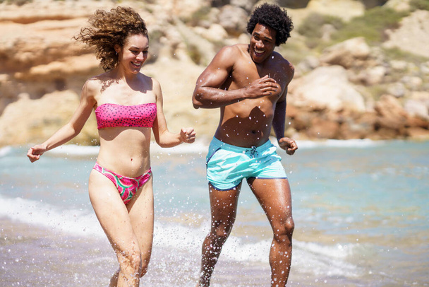Sonriente joven hombre afroamericano corriendo con compañera femenina en salpicaduras de agua y mirando hacia otro lado mientras disfrutan de la diversión juntos en la orilla del mar durante el día soleado - Foto, Imagen