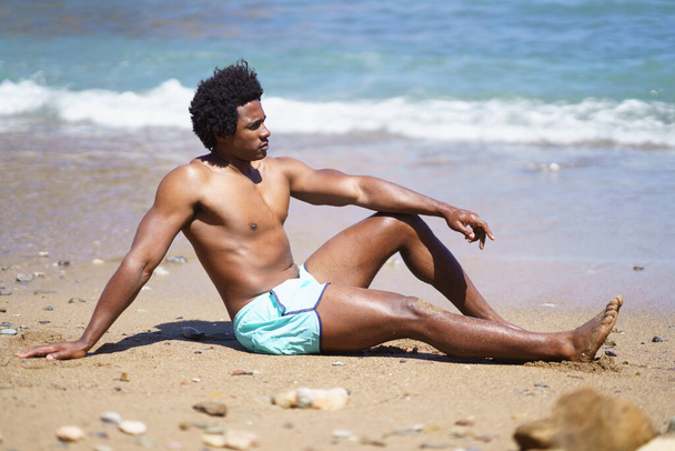 Full body side view van Afro-Amerikaanse man ontspannen op zandstrand met arm op knie, terwijl weg te kijken, en leunend op de hand in de buurt van golvende zee - Foto, afbeelding