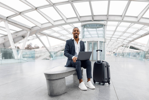 Чёрный молодой бизнесмен с ноутбуком, сидящий на скамейке в аэропорту, симпатичный афроамериканец тысячелетия в костюме, ожидающий полета в терминальном зале, наслаждающийся деловой поездкой, копировальным пространством - Фото, изображение