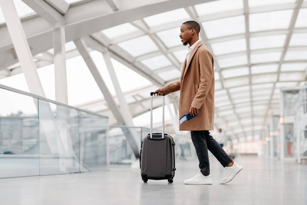 スタイリッシュなコートを着たハンサムな黒人男性空港でスーツケースを着て歩く、若いアフリカ系アメリカ人男性ターミナルホールを通過し、出発ゲートに行き、飛行機で一人旅、サイドビュー - 写真・画像