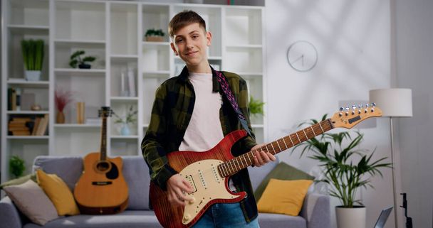 Caucasian teen boy dancing and playing on guitar and listening to the music in the living room. Happy and crazy, having fun. - Foto, Imagem