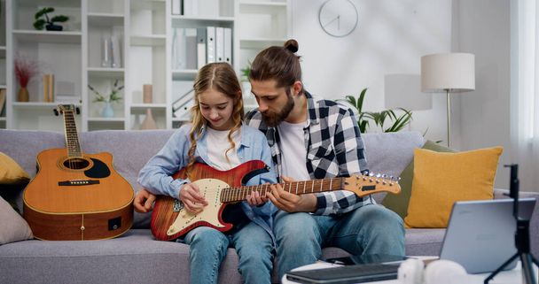 Father and daughter teaching, playing guitar and learning musical notes at home. Skilled talented personal tutor teaches his little student to be good at playing guitar. - Foto, Bild