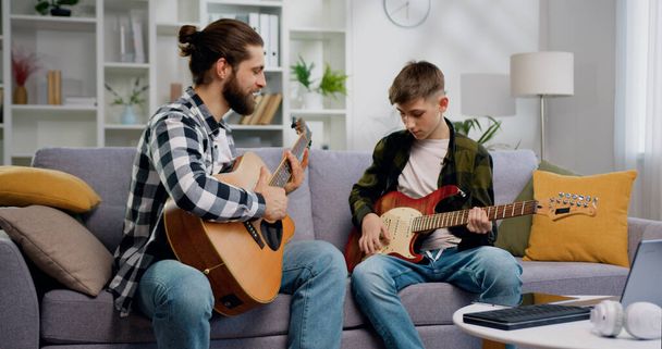 Father and son play guitar at home. Dad teaches child to play musical instruments. Spending happy time together. People and music concept. - 写真・画像