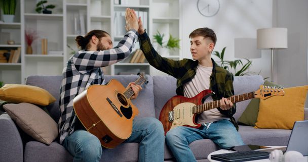 Creatieve tiener speelt gitaar terwijl de mens thuis kijkt en les geeft in het delen van ervaringen. Tiener jongen die gitaar leert spelen. Portret van een gelukkige vader en zoon. - Foto, afbeelding