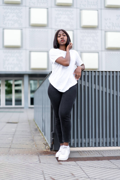 Chic and stylish: A portrait of a young black woman in a white tee and black pants against a grey building - Фото, изображение