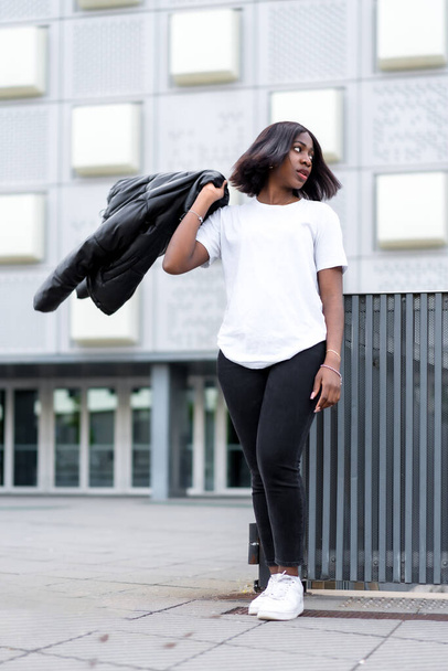 Urban cool: A portrait of a young black woman in a fashionable white t-shirt and black pants, posing in front of a sleek, cityscape backdrop - 写真・画像