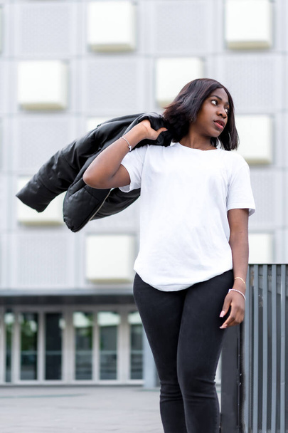 Simple yet stylish: A portrait of a young black woman in a minimalist white t-shirt and black pants, set against a sleek, modern building - Foto, imagen