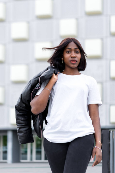 Fashionable city vibes: A young black woman strikes a pose in a stylish white tee and black pants, with a grey building as a chic urban backdrop - Photo, image
