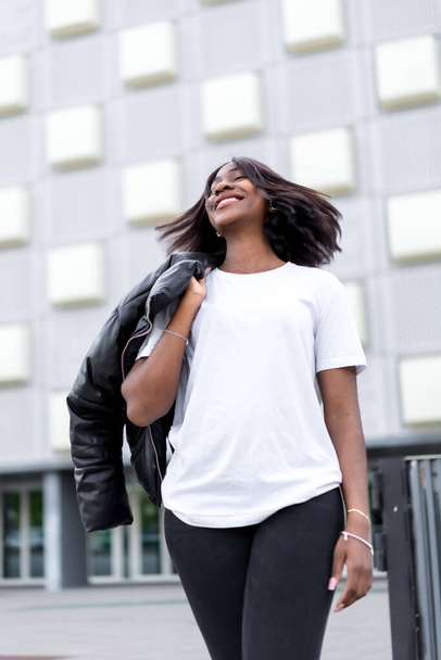 Minimalist chic: A portrait of a young black woman in a sleek white t-shirt and black pants, set against the backdrop of a contemporary grey building - Foto, Imagem