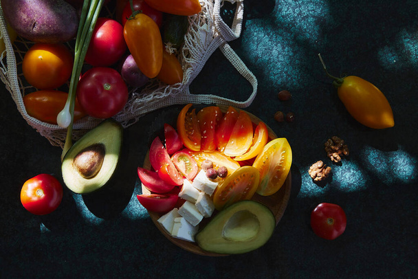 Здоровый сезонный салат с овощами, сыром фета. Лук, желтый, красные помидоры и авокадо, орехи в экологически чистой сумке под модными твёрдыми тенями с отражающим фоном. Здоровое питание - Фото, изображение