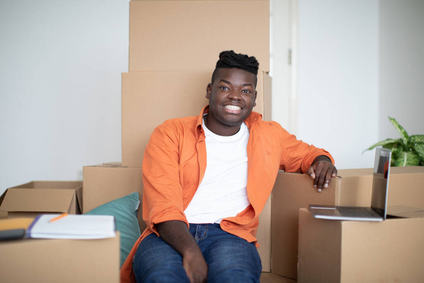 Homem negro sentado entre caixas de papelão e usando laptop depois de se mudar para casa, feliz jovem afro-americano masculino fazendo Checklist e compras on-line no computador enquanto se desloca para novo apartamento - Foto, Imagem