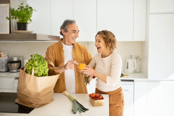 Iloinen vanhempi pari saapuu supermarketista ruokakassin kanssa ja purkaa tavaroita keittiössä, iäkkäät puolisot nauttivat elämästä, viettävät aikaa auttaen toisiaan. - Valokuva, kuva