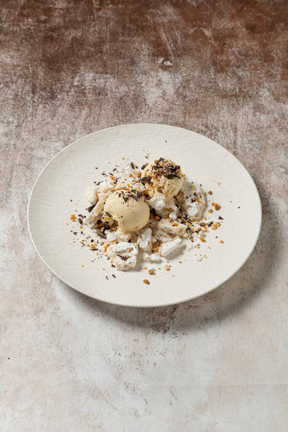 Two scoops of coffee flavored ice cream in a bowl with meringue, chocolate chips and caramel with hazelnuts. The dessert lies on a white ceramic plate on a light coffee-colored background. - Photo, Image