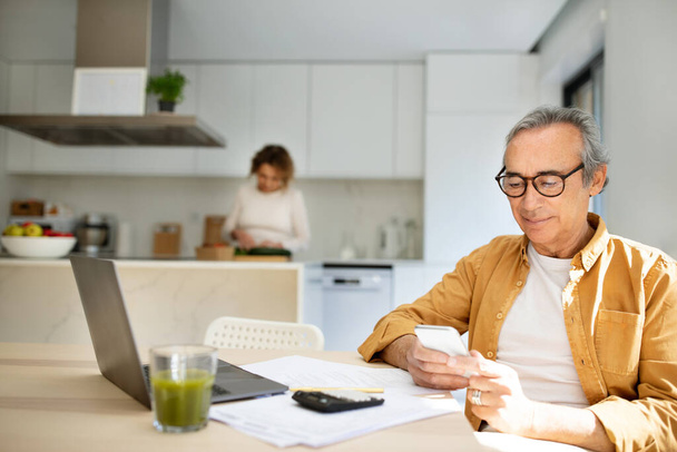 Senior man using smartphone, sitting at table with laptop in kitchen while his wife cooking lunch, surfing internet or messaging on mobile phone while waiting meal - Photo, Image