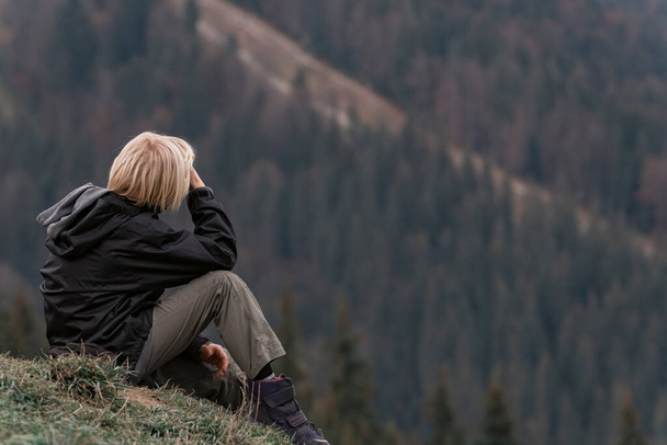 Το ξανθό αγόρι κάθεται στην πλαγιά του λόφου και κοιτάζει την απόσταση. Πλευρική άποψη για το αγόρι στα βουνά. Διακοπές με παιδιά. - Φωτογραφία, εικόνα