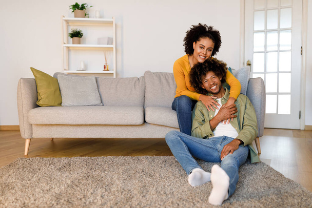 Milující africký americký pár pózování objímání v moderním obývacím pokoji, sedí na gauči a usmívá se na kameru doma. Spokojenost a pohodlí. Volné místo pro textovou reklamu - Fotografie, Obrázek
