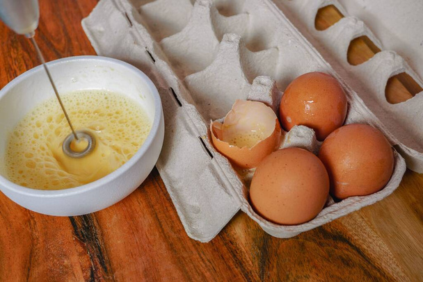 beating an egg in a bowl with a small mixer with an egg cup at the bottom on a wooden table - Foto, Imagen