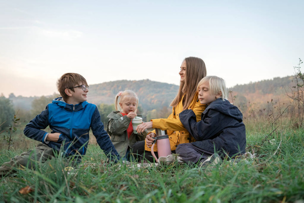 Happy mom and three kids have fun at picnic in fall. Two boys and girl with mother drink hot drink on mountains background. - Photo, Image